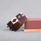 A brown polo leather belt with cream, blue, and red stitching, and a classic antique buckle, displayed alongside a gray object and two pencils.